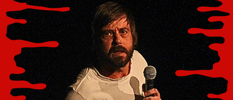 A pensive, gremliny, man stands in the dark with a microphone