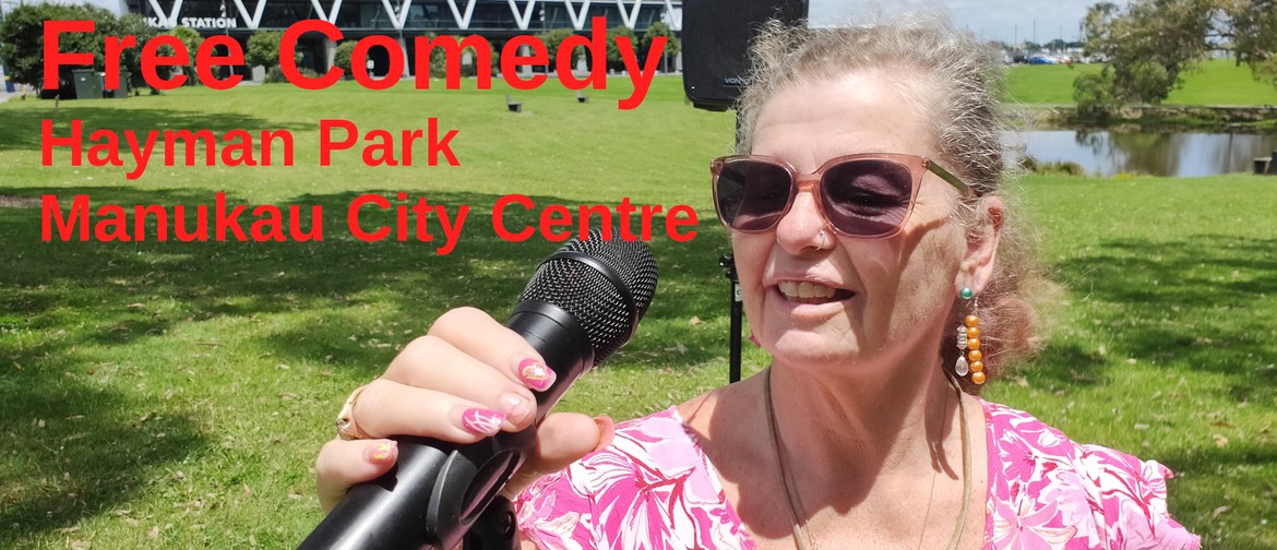 A comedian on a grassed park in front of the Manukau Railway station holds a microphone ready to tell jokes.