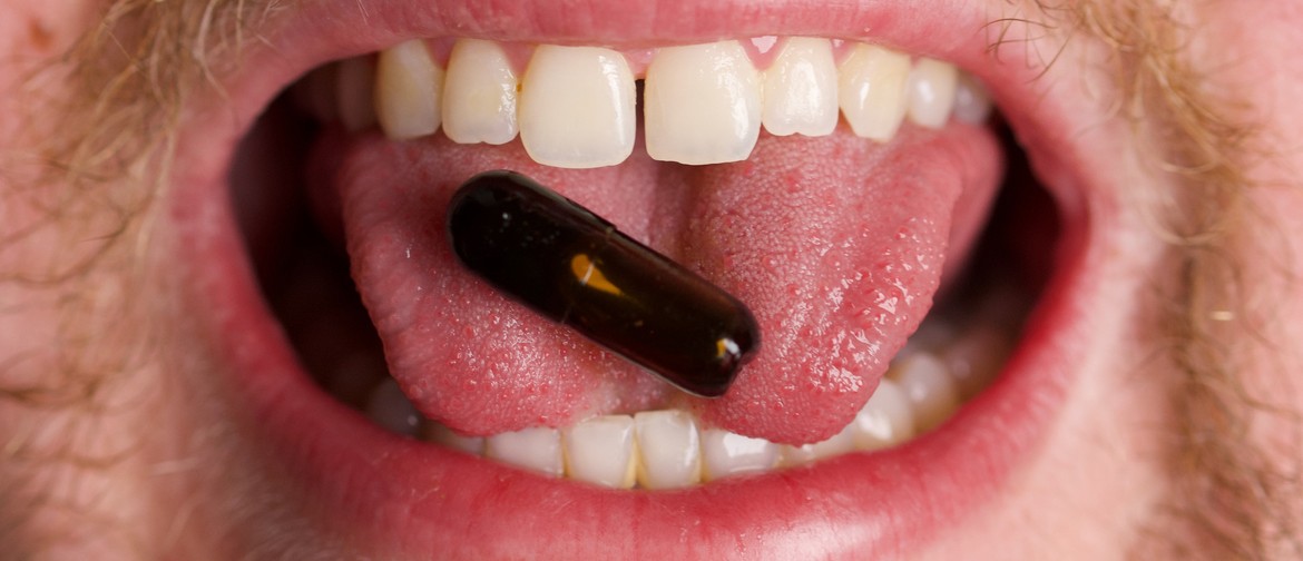 A close up image of an open mouth. On the tip of the tongue there is a black pill.