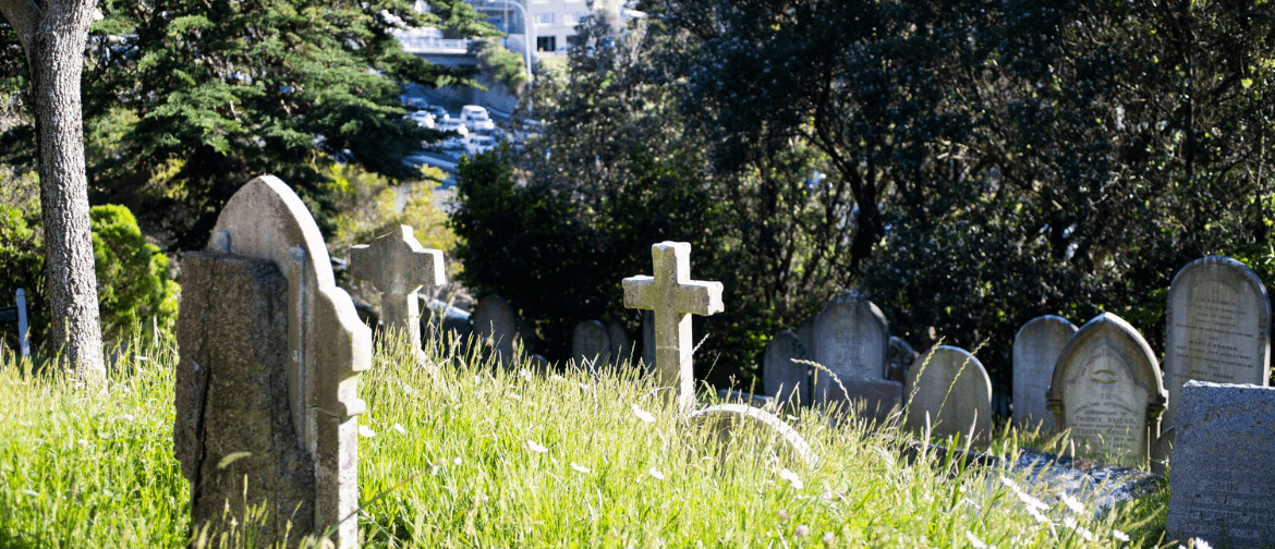 A landscape veiw of the cemetery with a headstone in the shape of a cross in the middle. Trees frame the image and the grass is long with meadow flowers. 