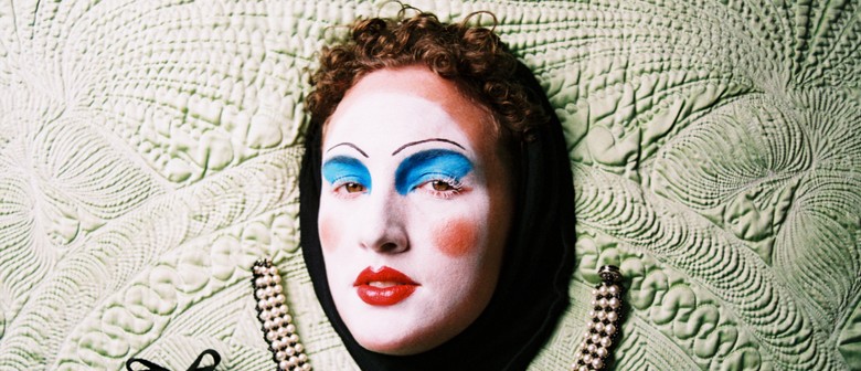 A face with clown-like makeup faces outward from a wall of textured cloth, looking pensive.