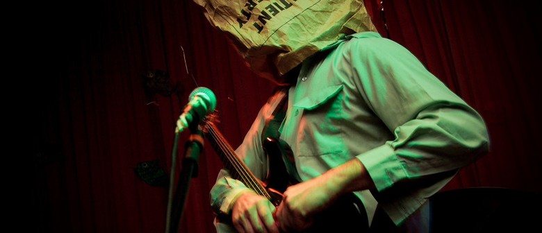 Surfing USSR Go Exotica! Musician stands at microphone in a dark venue with a paper bag over their head.