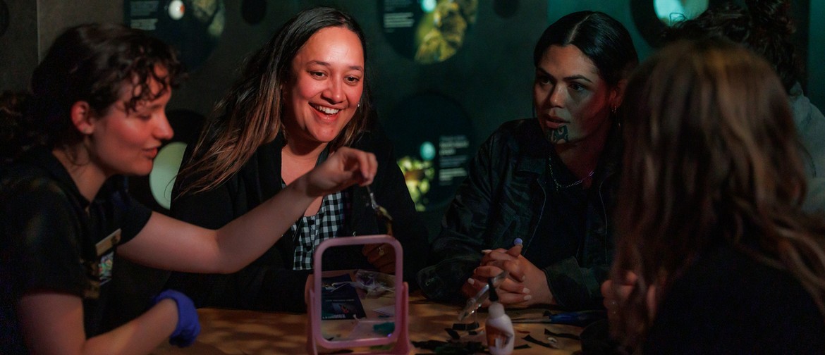 After Dark: Mātahi o Rongo A photo of 5 adults sit around a table in low lighting. The facilitator leads an activity demonstrating a creative action with a mirror. The 2 participants facing the camera are smile