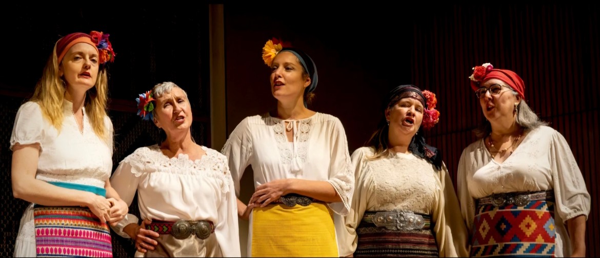 Medenki of The Medena Ensemble 5 women with stand on a darkened stage in various poses singing. They are dressed I whites and creams and have brightly colored scarves and flowers on their heads.